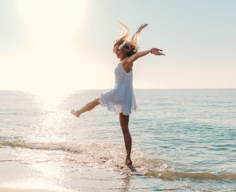 young attractive happy woman dancing turning around by sea beach sunny summer fashion style in white dress vacation